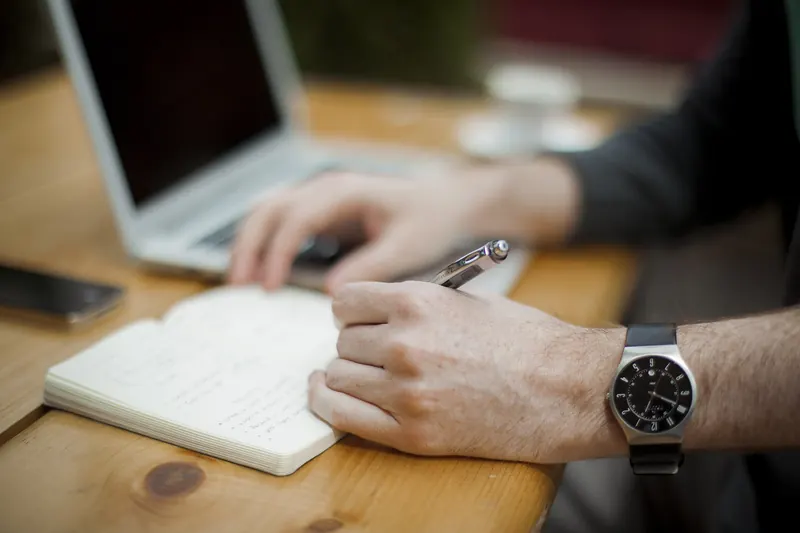 Person writing a list in a notebook beside a laptop at a wooden table