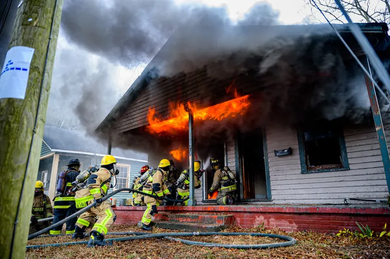 Firefighters working a hose line as flames burn through the front porch of a wood-frame house