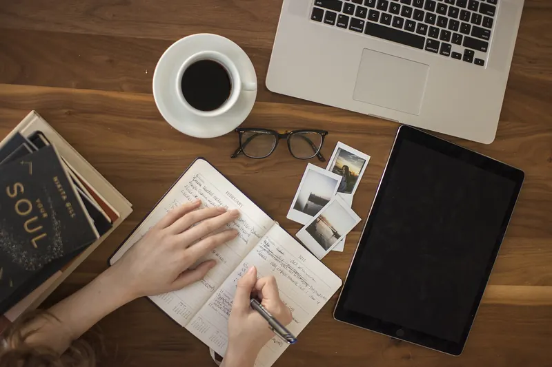 Overhead view of a desk with a notebook being filled in, laptop, tablet, glasses and coffee
