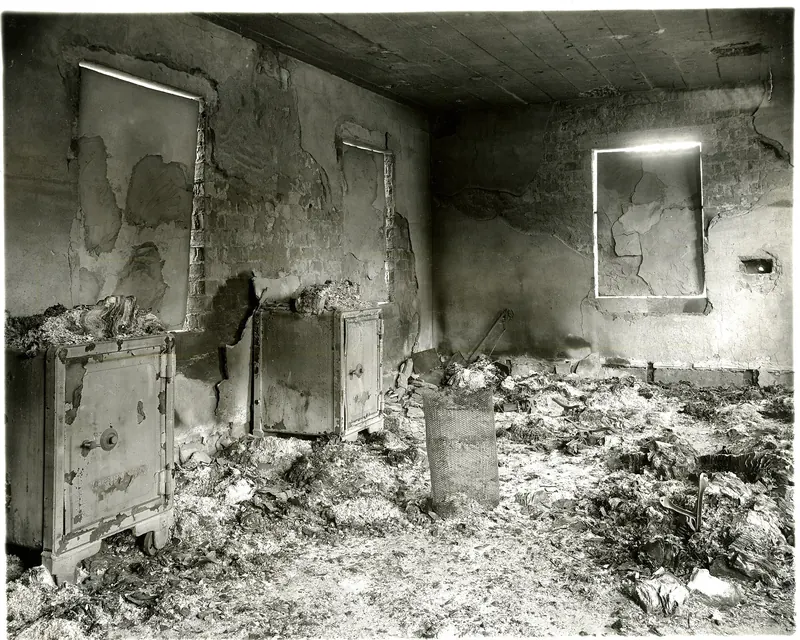 Black-and-white view of a fire-gutted room with soot-darkened ceiling, cracked plaster and burned debris